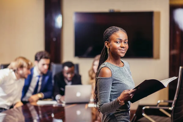 Empresária africana olha para câmera na sala de reuniões com colegas de fundo — Fotografia de Stock