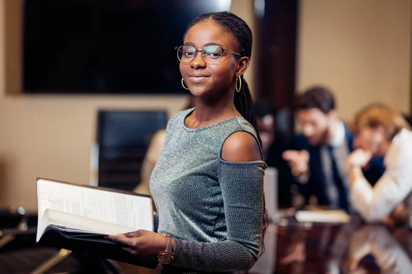 Empresária africana olha para câmera na sala de reuniões com colegas de fundo — Fotografia de Stock