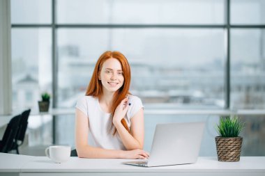 Güzel kızıl kız dizüstü bilgisayarda çalışıyor ve kameraya bakıyor.