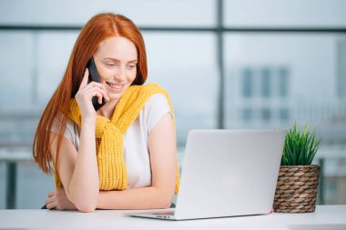 İş kadını masada oturuyor, dizüstü bilgisayara bakıyor ve akıllı telefon kullanıyor.