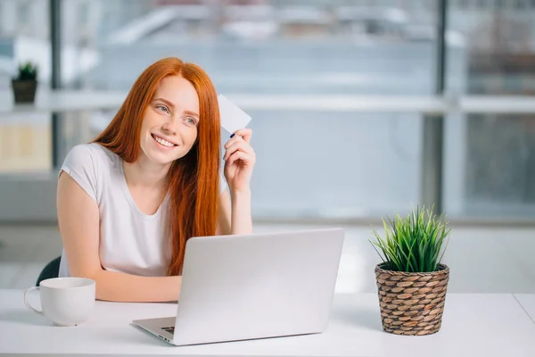 Happy redhead shopper deciding what to buy on line holding a credit card sit — Stok Foto