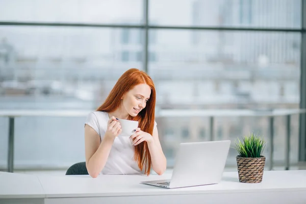 Mutlu gülümseyen kızıl saçlı kadın laptopla çalışıyor ve kahve içiyor. — Stok fotoğraf