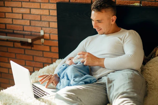 Hombre alimentando pequeño bebé mientras está acostado en la cama y trabajando en el ordenador portátil —  Fotos de Stock