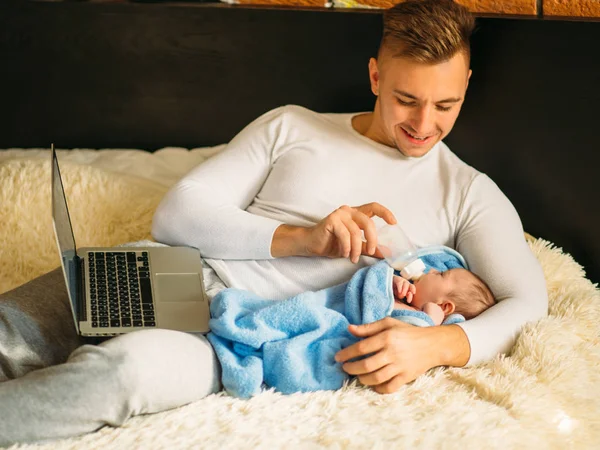 Man feeding little baby while lying on bed and working on laptop — Stock Photo, Image