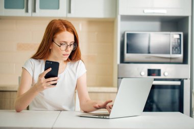 mutfakta oturuyor ve dizüstü bilgisayar kullanırken telefonu tutan kadın