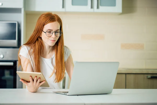 Redhead using the tablet pc and laptop in kitchen — Stock Photo, Image