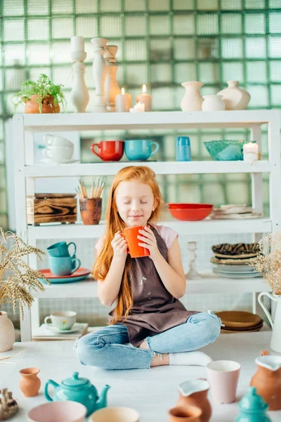 Рыжая девушка сидит в мастерской глины студия с большим количеством чашки — стоковое фото