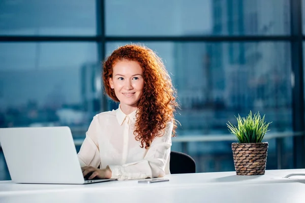 Rothaarige Geschäftsfrau sitzt am hellen modernen Arbeitsplatz und tippt am Laptop — Stockfoto