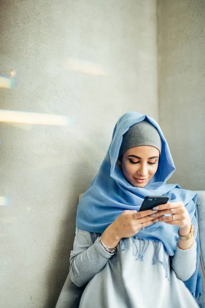 Potret wanita muslim yang bahagia menggunakan ponsel saat duduk di sofa — Stok Foto
