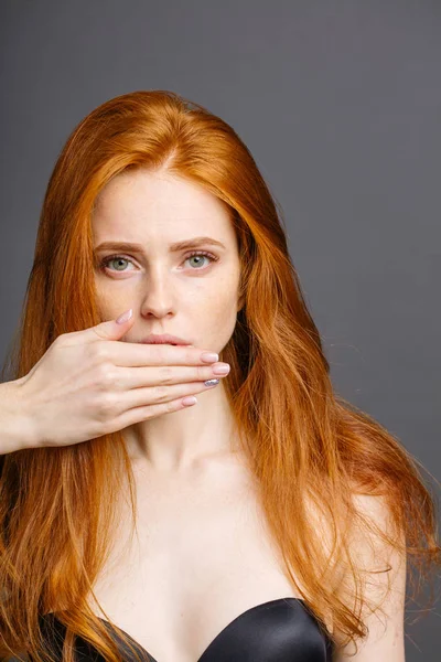 Ruiva mulher cobrindo boca com a mão e olhando para a câmera no fundo cinza — Fotografia de Stock