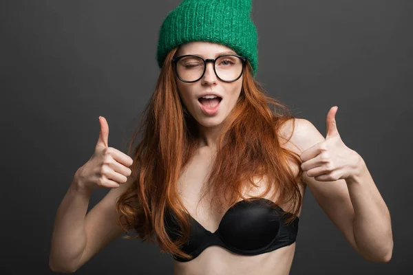 Рыжая женщина в черном лифчике, очках и зеленой вязаной шляпе показывает два больших пальца вверх — стоковое фото