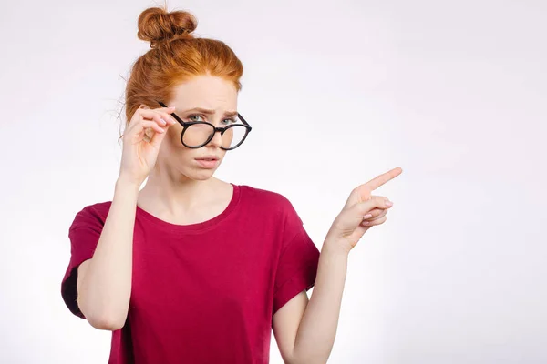 Pelirroja en gafas apuntando con el dedo al espacio de copia sobre fondo blanco —  Fotos de Stock