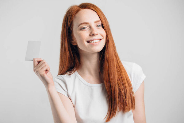 Рыжая девушка держит кредитку и улыбается на белом фоне
.