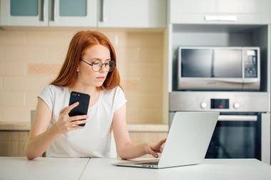 mutfakta oturuyor ve dizüstü bilgisayar kullanırken telefonu tutan kadın