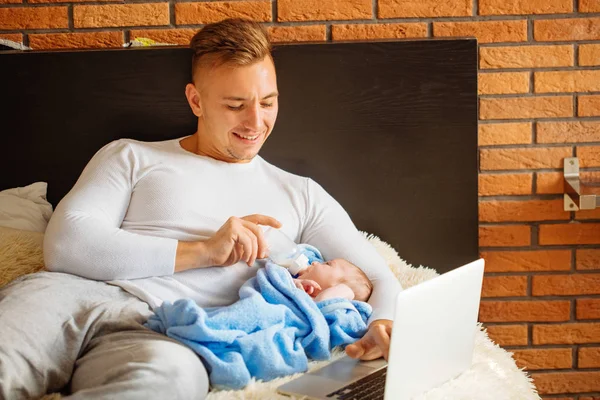 Man feeding little baby while lying on bed and working on laptop — Stock Photo, Image