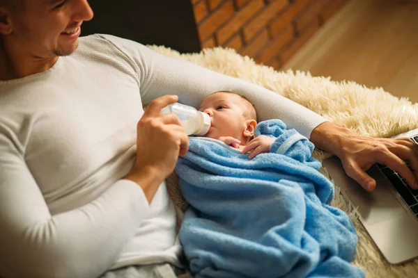 Hombre alimentando pequeño bebé mientras está acostado en la cama y trabajando en el ordenador portátil — Foto de Stock