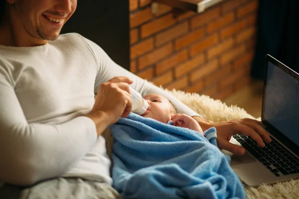 Uomo nutrire piccolo bambino mentre sdraiato sul letto e lavorare sul computer portatile — Foto Stock