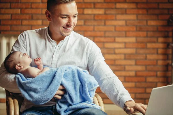 Mann arbeitet zu Hause am Schreibtisch mit Laptop, hält niedliche Babys in der Hand, schaut auf den Bildschirm — Stockfoto