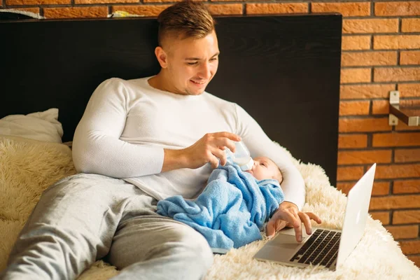 Człowiek karmienia małego dziecka podczas leżąc na łóżku i podczas pracy na laptopie — Zdjęcie stockowe