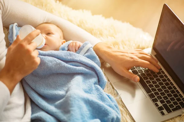 Muže, který krmí děťátko ležící na posteli a práci na notebooku — Stock fotografie