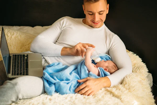 Homem alimentando pequeno bebê enquanto deitado na cama e trabalhando no laptop — Fotografia de Stock