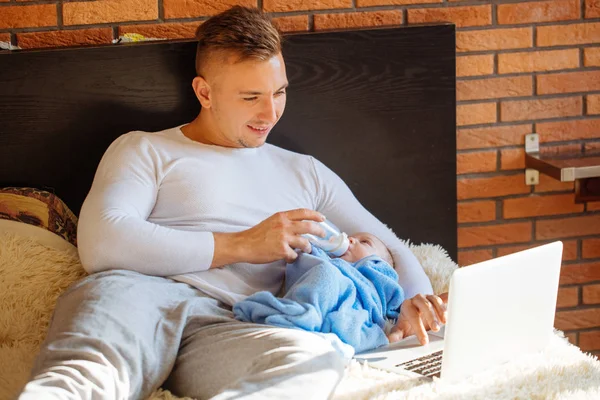Hombre alimentando pequeño bebé mientras está acostado en la cama y trabajando en el ordenador portátil —  Fotos de Stock