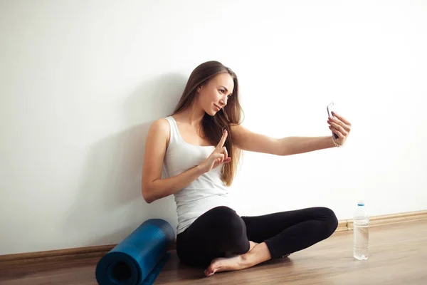 Женщина сидит на полу и делает селфи на смартфоне в тренажерном зале — стоковое фото