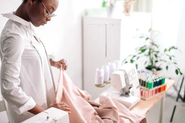 Mulher africana costureira olhando para tecidos rosa e de pé na oficina — Fotografia de Stock