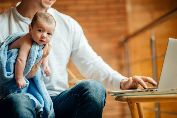 Muž, který pracuje u stolu doma s notebookem, držel roztomilý babys, při pohledu na obrazovku — Stock fotografie
