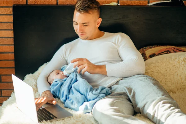 Hombre alimentando pequeño bebé mientras está acostado en la cama y trabajando en el ordenador portátil — Foto de Stock