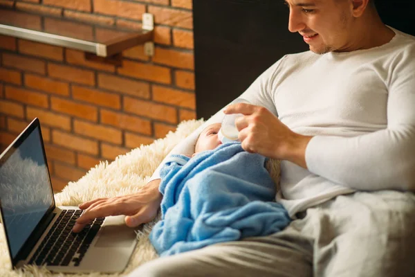 Muže, který krmí děťátko ležící na posteli a práci na notebooku — Stock fotografie