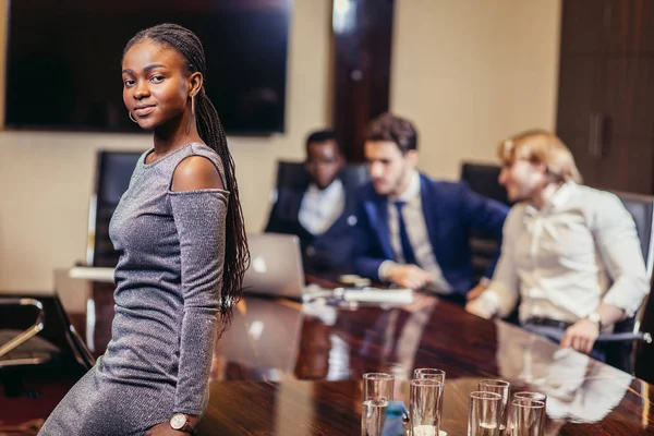 Empresária africana olha para câmera na sala de reuniões com colegas de fundo — Fotografia de Stock
