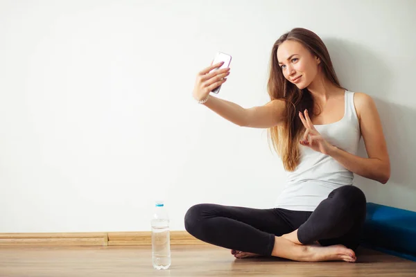 Женщина сидит на полу и делает селфи на смартфоне в тренажерном зале — стоковое фото