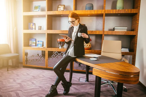 woman drinking coffee and reading her tablet while standing in office