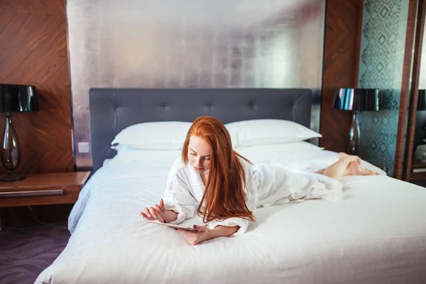 Όμορφη γυναίκα, φορώντας το μπουρνούζι και χρησιμοποιώντας ψηφιακή δισκίο ενώ χαλαρώνετε στο κρεβάτι — Φωτογραφία Αρχείου