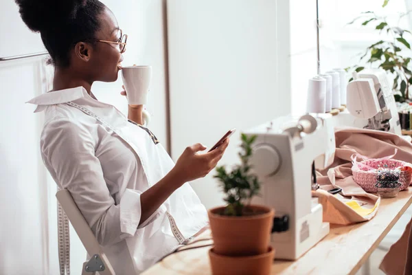 Vrolijke vrouw naaister in schort praten over telefoon en koffie drinken — Stockfoto