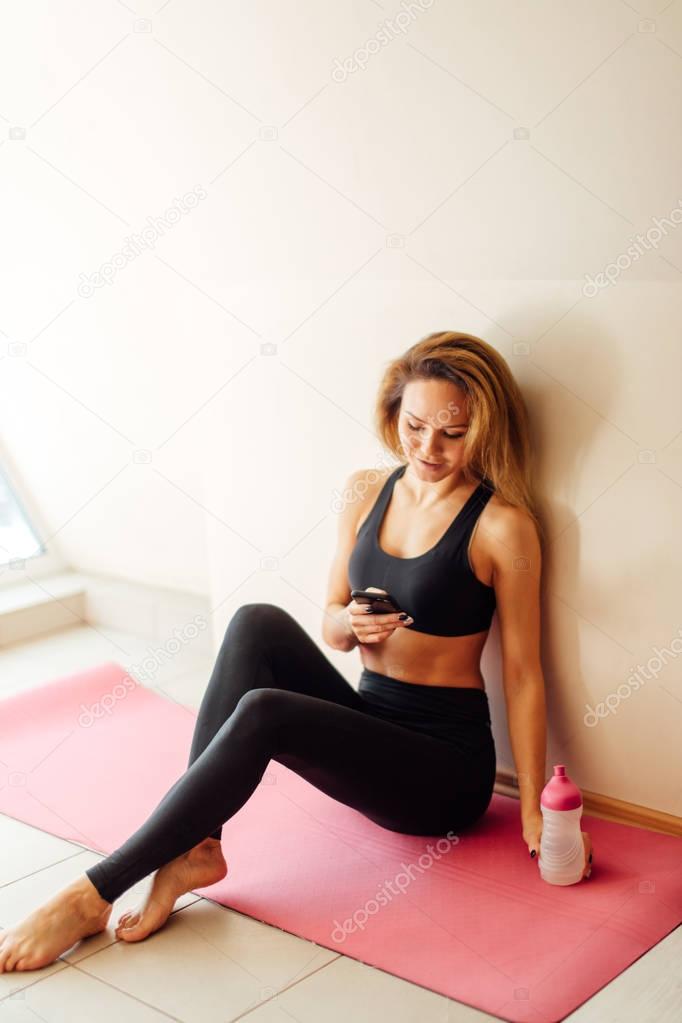 girl holding smart phone, resting on the white wooden floor after exercises
