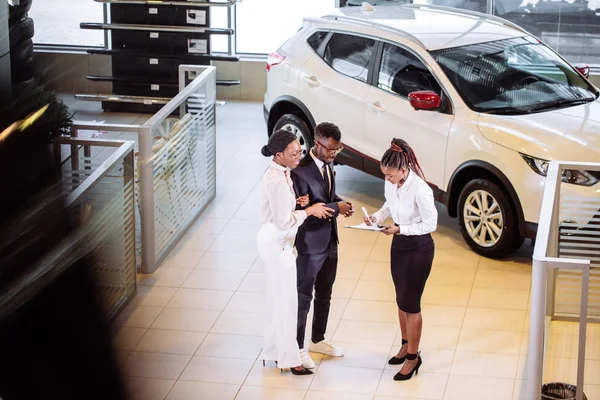 Car salesman standing at dealership telling about features of car to customers — Stock Photo, Image