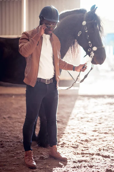 Africano hombre usando gafas de sol cerca negro caballo en hangar — Foto de Stock