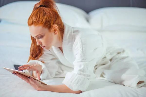 Vacker kvinna klädd i morgonrock och använda digitala surfplatta medan du kopplar av på säng — Stockfoto