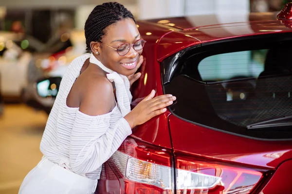 Sorridente donna africana abbracciando auto rossa al nuovo showroom auto — Foto Stock