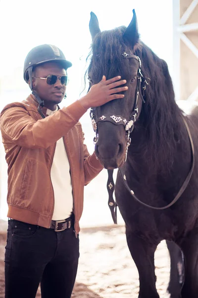 Africano hombre usando gafas de sol cerca negro caballo en hangar — Foto de Stock