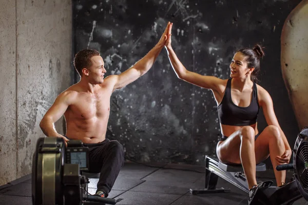 Man and woman giving high-five while doing exercises with rowing machine — Stock Photo, Image