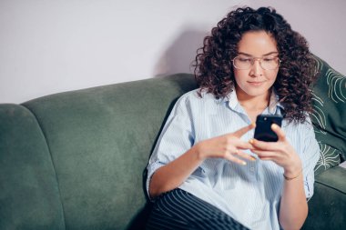 cropped image of adult with curly hair sitting on the grey sofa with phone clipart