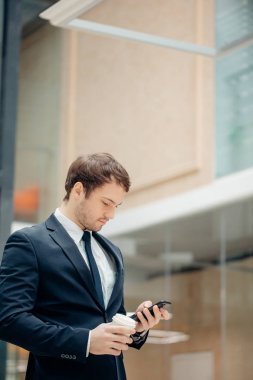 kahve fincanı tutarak ve koridorda yürürken akıllı telefonunu kullanarak kendine güvenen adam