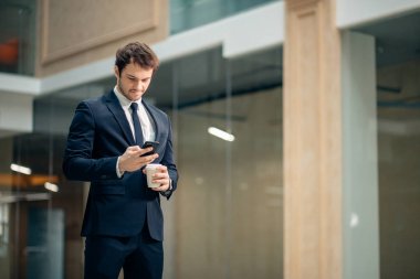 kahve fincanı tutarak ve koridorda yürürken akıllı telefonunu kullanarak kendine güvenen adam