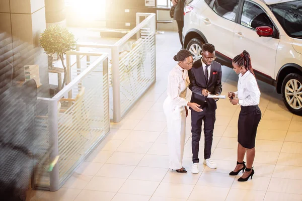 Car salesman standing at dealership telling about features of car to customers — Stock Photo, Image