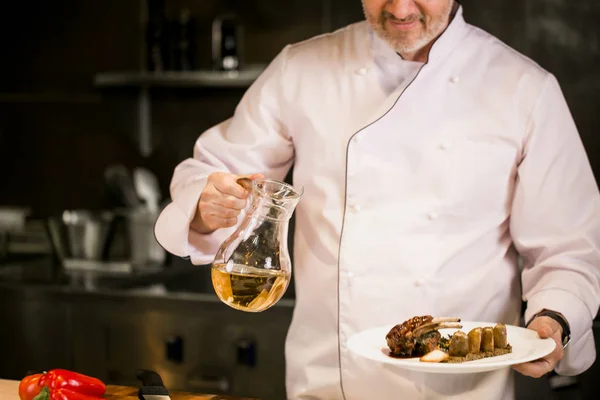 Viejo cocinero de cabeza en manteles blancos sosteniendo el frasco de aceite y plato con sabroso plato — Foto de Stock