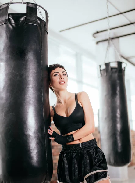 Hermosa mujer boxeador en gimnasio clothers al lado de gran saco de boxeo negro —  Fotos de Stock