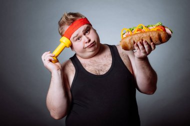 Şişman adam kameraya bakıyor, hamburger ve dambıl tutuyor.